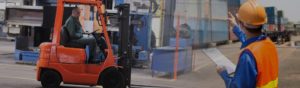 Worker drives safety through manufacturing truck on forklift and other worker with safety vest and hardhat directs traffic